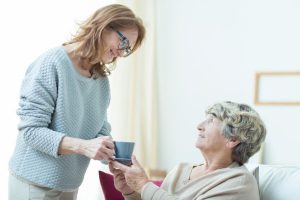 Middle-aged lady smiles as she hands a mug to an elderly woman sitting in bed